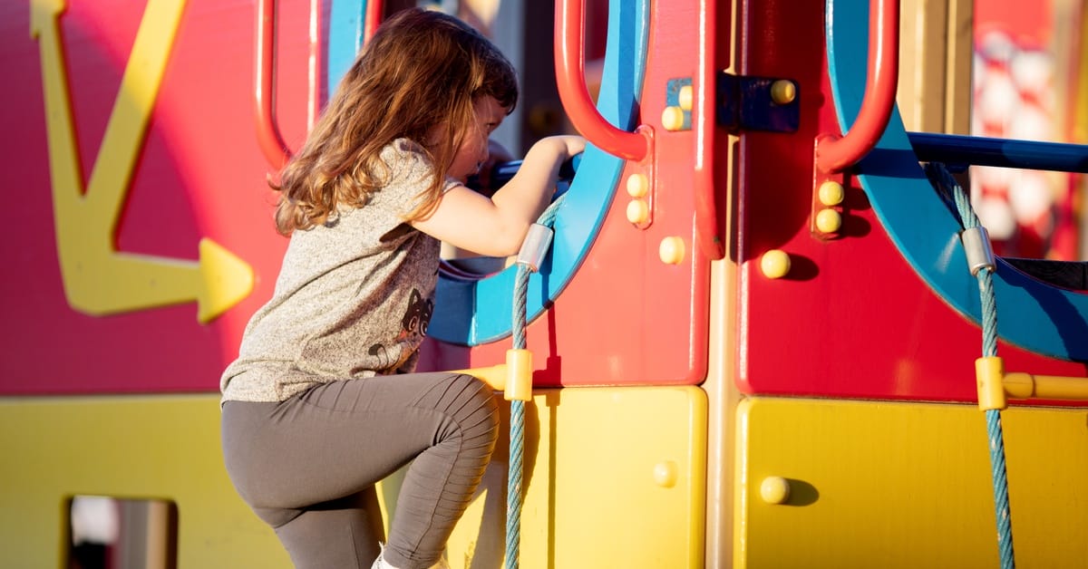 A little kid is playing on a colorful playground at the park. They're climbing up a roped ladder to the slide.