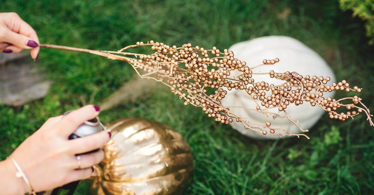 A close-up shows a person spray painting a floral element with gold paint. A white pumpkin and a gold pumpkin are in the back.