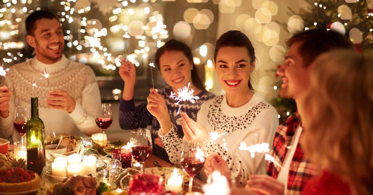 A group of people at a festive dinner table with food, wine, and candles. They hold sparklers, surrounded by lights.