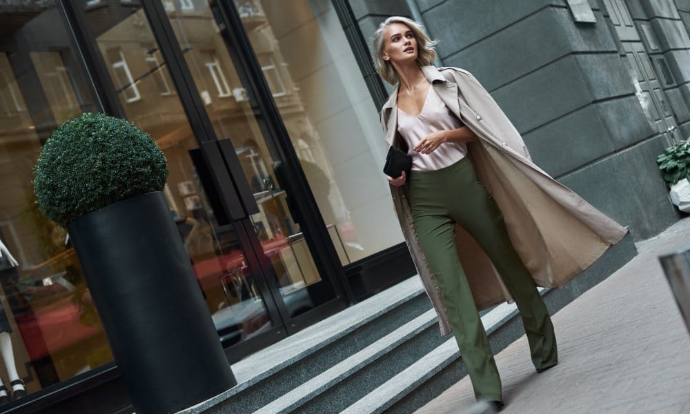 A blonde woman wearing dark green pants, an off-white blouse, and a trench coat walks along a brick walkway outdoors.