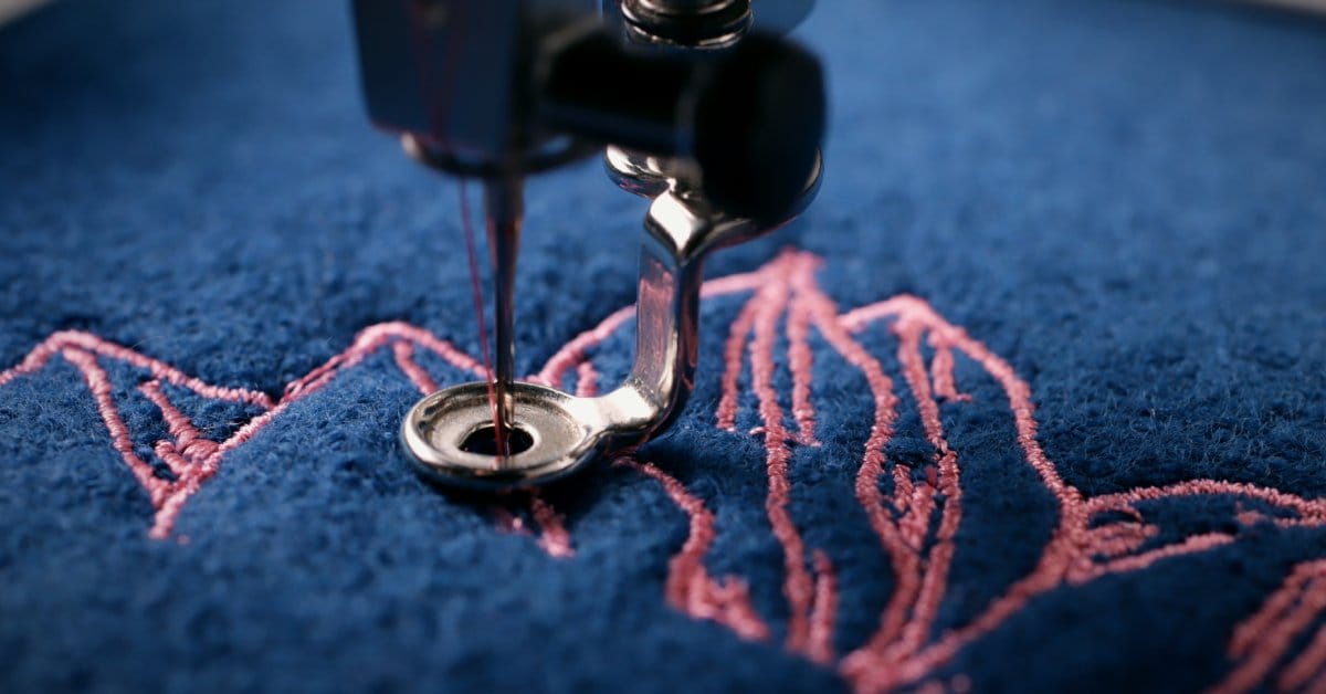 A machine embroidery needle stitches pink thread into a dark blue fabric. The design looks like petals or leaves.