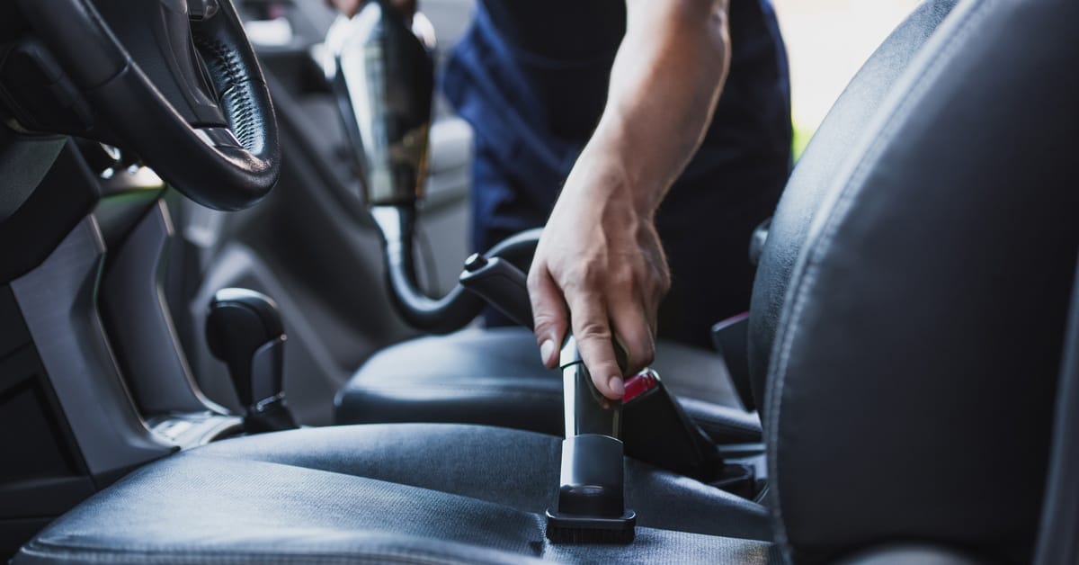 A person is pressing a vacuum nozzle against the seat of a car. The interior of the car is black and the person is standing outside the car.