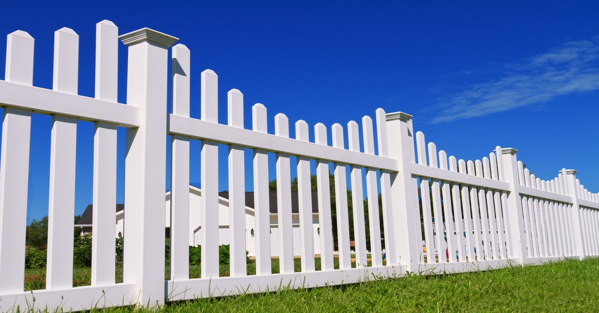 A close-up of a white vinyl fence in a residential neighborhood. The grass is bright green on a sunny day.