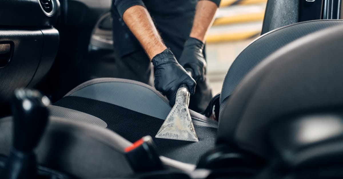 A man with black gloves and a black shirt with rolled up sleeves is using a vacuum to clean a car seat.