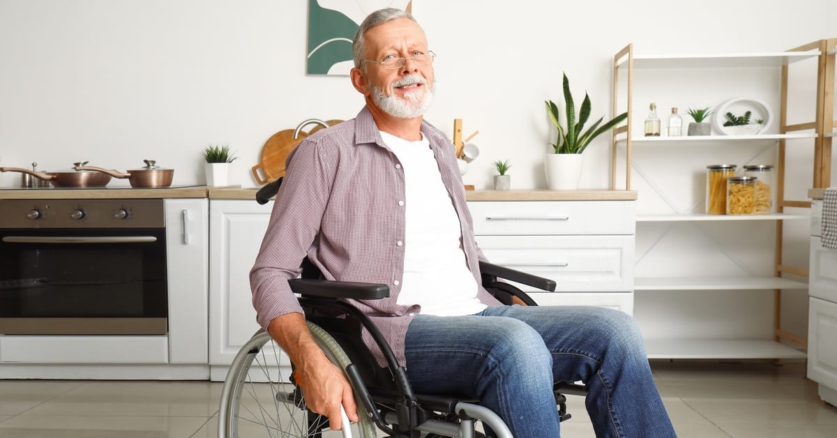 An elderly man in a wheelchair in the middle of his kitchen. Cabinets are white with cabinet pulls for easy access.