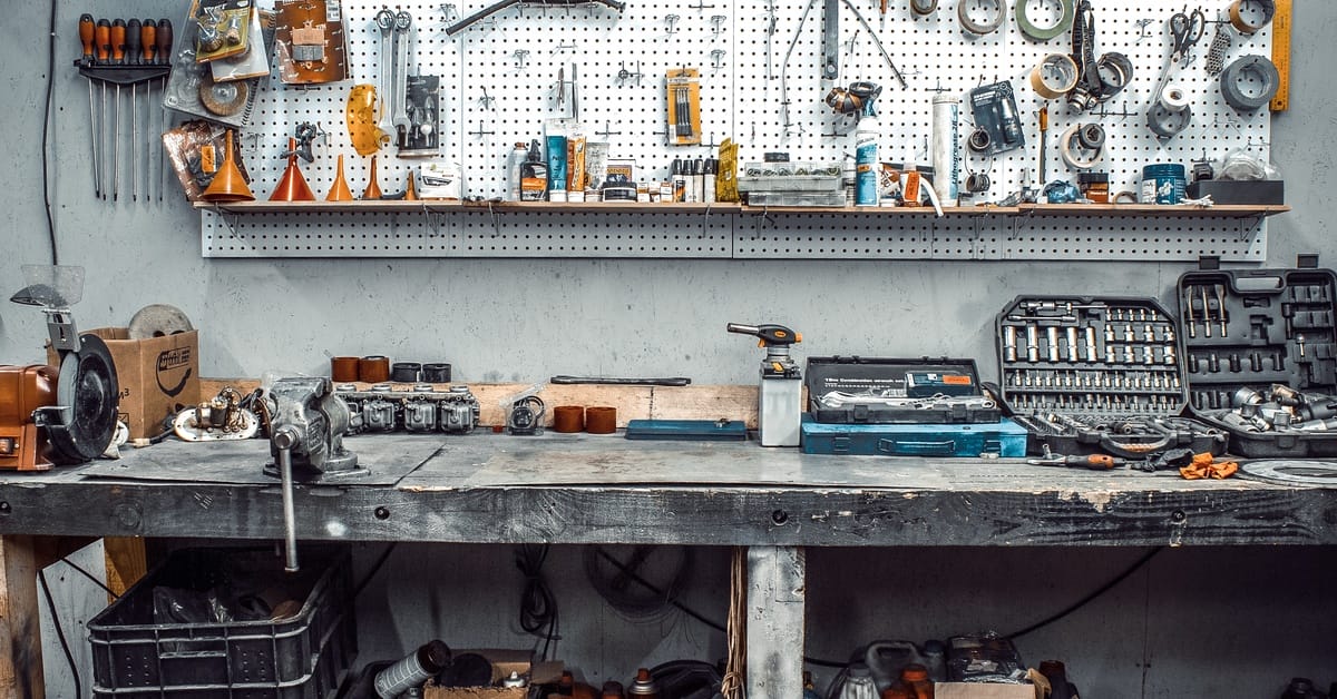 A wooden table with various tools strewn on the surface. There is a tool rack on the back wall.