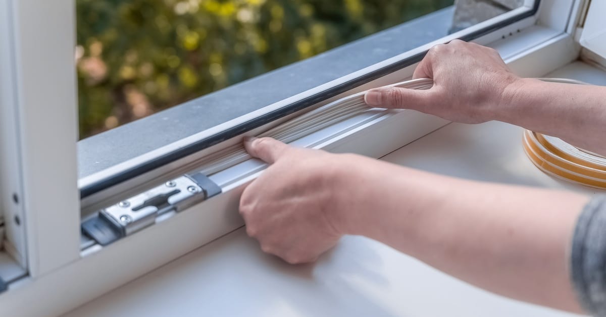 A person is applying a sealer to the edge of a window frame. There are green trees just outside the window.
